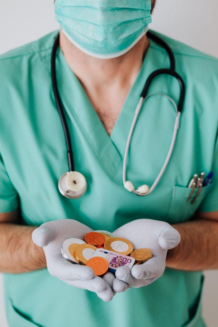 gallery-05 Anonymous male doctor wearing medical uniform mask and latex gloves holding coins and plastic euro while standing near white wall in clinic