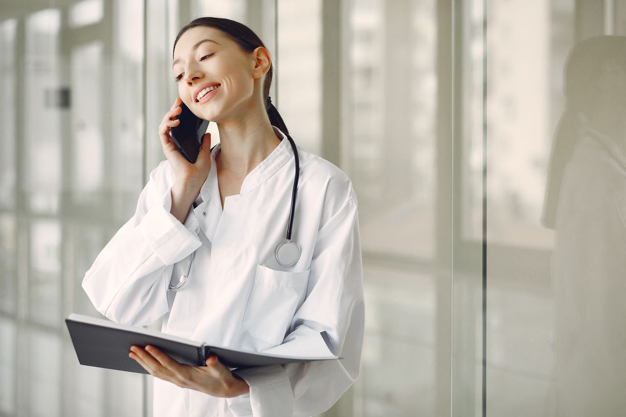 gallery-06 Smiling ethnic female doctor in medical uniform with stethoscope and notebook talking on smartphone while standing in glass hospital corridor