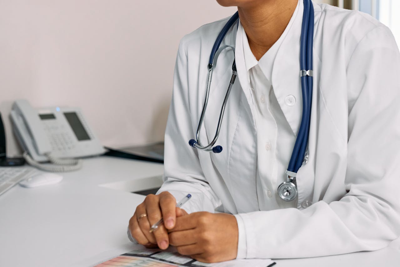 gallery-02 Doctor with stethoscope and white coat seated at a clinic desk.