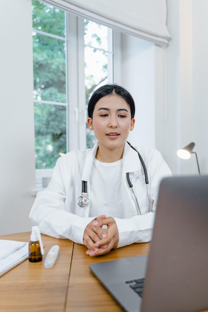 Asian female doctor in white coat using a laptop for online consultation from her office desk.