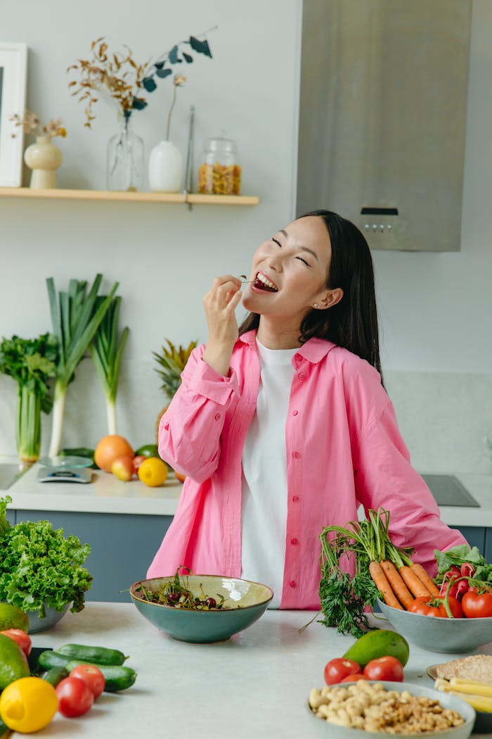 Asian woman in a pink shirt enjoying fresh vegetables in a modern kitchen setting.